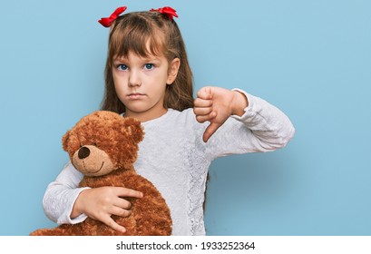 Little Caucasian Girl Kid Hugging Teddy Bear Stuffed Animal With Angry Face, Negative Sign Showing Dislike With Thumbs Down, Rejection Concept 