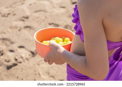 Little Caucasian Girl With Purple Bikini Eating Green Grapes Fruit As Dessert From An Orange Recipient While Standing In The Beach