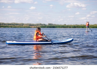 Little Caucasian girl sitting on paddle board alone holding oar in hands and looking at other sup boarders in orange life jacket. Active holidays. Inculcation of love for sports from childhood. - Shutterstock ID 2106571277