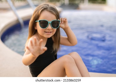 Little Caucasian Girl In Sunglasses And Black Bathing Suit Waving Hello, Sitting By The Pool In Summer. Little Cute Girl Relaxing In Swimming Pool. Summer Travel Hotel Vacation - Image