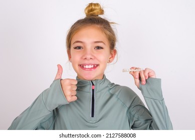 Little Caucasian Kid Girl With Hair Bun Wearing Technical Shirt Over White Background Holding An Invisible Braces Aligner And Rising Thumb Up, Recommending This New Treatment. Dental Healthcare 