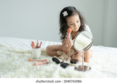 Little Caucasian Kid Girl Holding Pink Blush Brush To Apply Powder To Cheeks And Face Looking At Mirror With Joy On Bed At Home. Cute Child Playing Makeup Cosmetics With Bear Doll. Copy Space