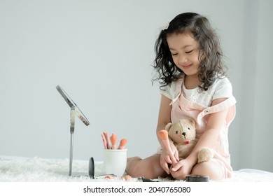 Little Caucasian Kid Girl Playing Makeup Cosmetics Sitting With Bear Doll. Cute Child Smiling And Holding Pink Blush Brush With Joy And Happiness On Bed At Home. Copy Space