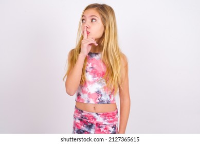 Little Caucasian Kid Girl Wearing Sport Clothing Over White Background Silence Gesture Keeps Index Finger To Lips Makes Hush Sign. Asks Not To Share Secret