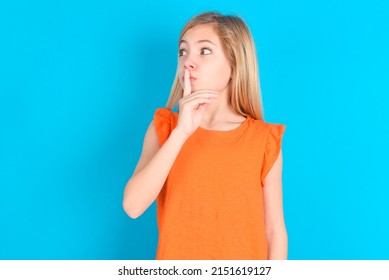 Little Caucasian Kid Girl Wearing Orange T-shirt Over Blue Background Makes Silence Gesture, Keeps Index Finger To Lips Makes Hush Sign. Asks Not To Share Secret.