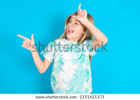 little caucasian kid girl wearing trendy T-shirt over blue background showing loser sign and pointing at empty space