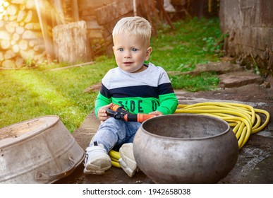 Little Caucasian Toddler Sitting On The Ground Outside In The Rural Ambience And Holding A Watering Hose.