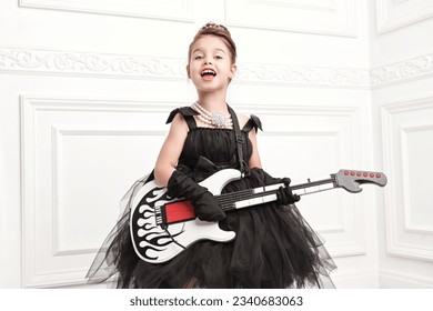 Little celebrity girl. A funny pretty girl in a luxury evening dress and with an evening hairstyle plays the electric guitar and sings. Pop and rock music culture. - Shutterstock ID 2340683063