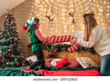 Little cheerful boy in an elf hat jumps on the bed holding his mother's hands