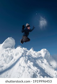 Little cheerful boy in sportswear jumps from a white snowy mountain against a blue sky.