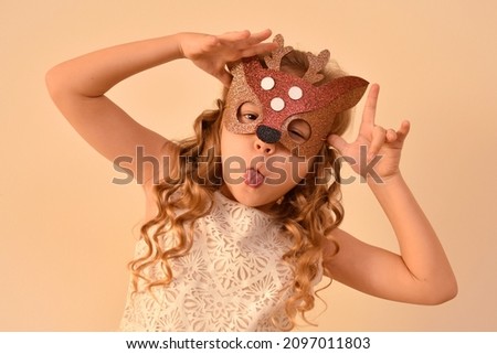 A little cheerful funny cute girl in a carnival masquerade mask of a handmade foamiran deer is having fun grimacing at the camera on a beige background.