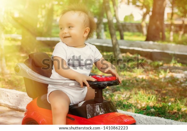 Little cheerful\
infant in white shirt sitting half turned back on red push car in\
park or garden in sun light. Happy active childhood and family\
vacation concept. Toned\
image