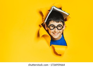 A little cheerful nerd boy in black glasses and an open book on his head break through a yellow colored paper wall. Sidebar for advertisements, events. Back to school and good luck.