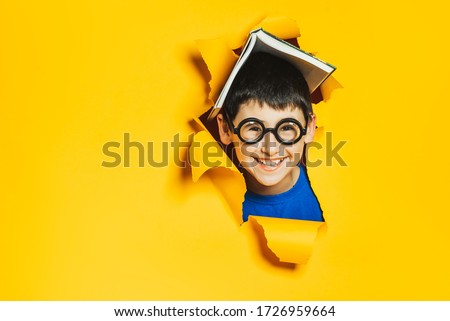 A little cheerful nerd boy in black glasses and an open book on his head break through a yellow colored paper wall. Sidebar for advertisements, events. Back to school and good luck.