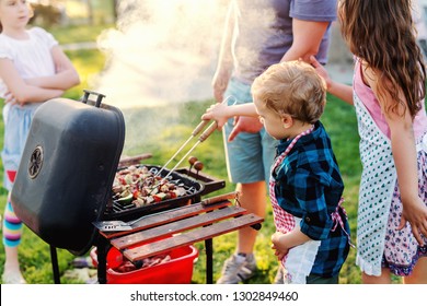 Little Chef With Apron Grilling Meat And Vegetables On Sticks. Next To Him His Cousins. Family Gathering Concept.