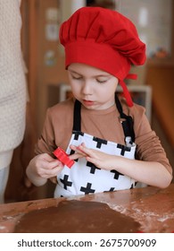 Little chef in red hat using cookie cutter for gingerbread dough before Christmas, holiday tradition at home. Copy space.