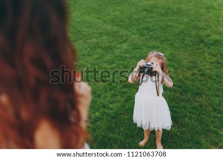 Little child baby girl in light dress take picture of mom on retro vintage photo camera on green grass in park. Mother, little kid daughter. Mother's Day, love family, parenthood, childhood concept
