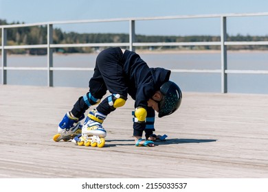 A little child in blue-yellow-white roller skates, helmet and protective gear learns to rollerblade falling on his hands. A walk on the sea wooden pier. Sunny summer day