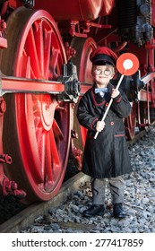 Little Child Boy As Nostalgic Railroad Conductor With Cap And Signaling Disk Beside Large Wheels Of A Steam Locomotive/Little Train Conductor Boy