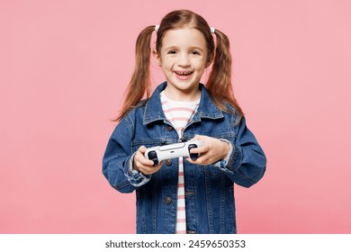 Little child cute kid girl 7-8 years old wears denim shirt hold in hand play pc game with joystick console isolated on plain pastel light pink background. Mother's Day love family lifestyle concept
