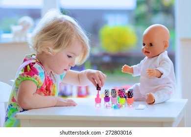 Little Child, Cute Toddler Girl Having Fun Playing At Home With Colorful Nail Polish Doing Manicure And Painting Nails To Her Doll