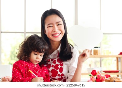 Little Child Daughter Girl Sitting On Mom's Lap, Kid Preparing Handmade Card To Celebrate Mothers Day While Happy Smiling Beautiful Young Asian Mom Holding Blank Heart Shaped Paper For Message.