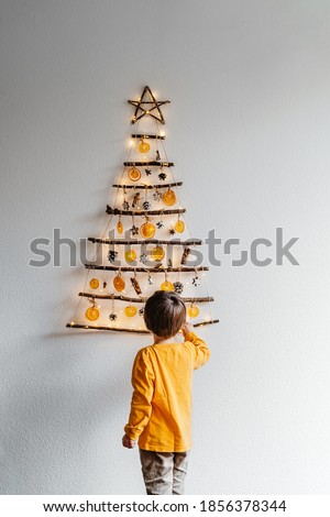 Little child decorating handmade craft Christmas tree made from sticks and natural materials hanging on wall. Sustainable Christmas, zero waste, plastic free, eco friendly.