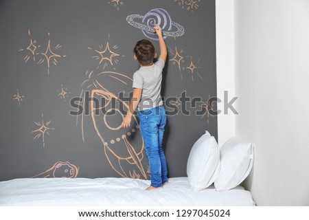 Little child drawing rocket with chalk on wall in bedroom