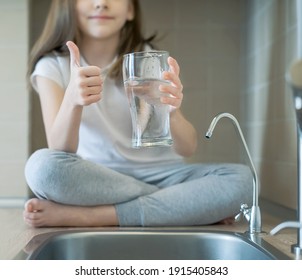 Little Child Drinking From Water Tap Or Faucet In Kitchen. Young Girl Showing Thumbs Up Sign And Holding A Transparent Glass. Kid Recommend Fresh Water. Water Quality Check Concept. World Water Day