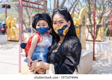 Little Child Girl And Asian Mother Wearing Medical Protective Mask And Holding Her Daughter In A Crowded Public Place In Park Or Tourist. Happy Baby And Mom Family Playing Outside Covid-19 Pandemic.