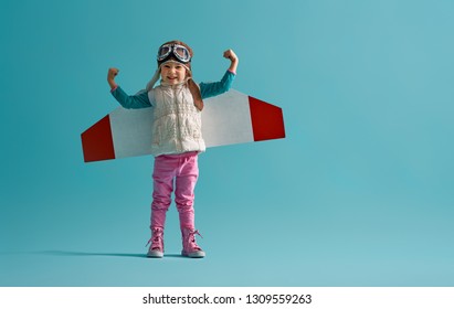 Little Child Girl In An Astronaut Costume Is Playing And Dreaming Of Becoming A Spaceman. Portrait Of Funny Kid On A Background Of Bright Blue Wall.