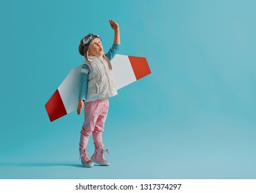 Little Child Girl In An Astronaut Costume Is Playing And Dreaming Of Becoming A Spaceman. Portrait Of Funny Kid On A Background Of Bright Blue Wall.