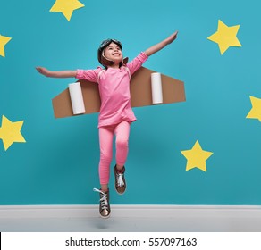 Little Child Girl In An Astronaut Costume Is Playing And Dreaming Of Becoming A Spaceman. Portrait Of Funny Kid On A Background Of Bright Blue Wall With Yellow Stars.