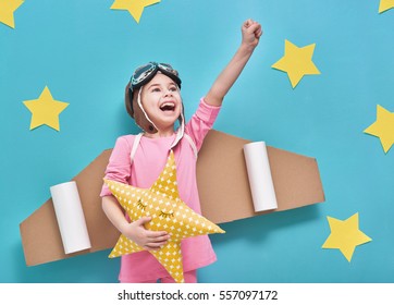 Little Child Girl In An Astronaut Costume Is Playing And Dreaming Of Becoming A Spaceman. Portrait Of Funny Kid On A Background Of Bright Blue Wall With Yellow Stars.