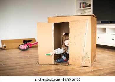 Little child girl looking out of a cardboard playhouse while moving in a new house. Lockdown activity idea.