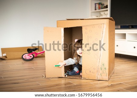 Little child girl looking out of a cardboard playhouse while moving in a new house. Lockdown activity idea.