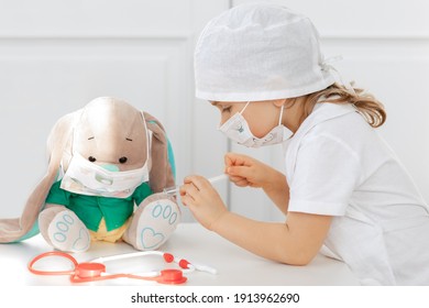 Little Child Girl Makes Injection To Toy. Cute Child With Medical Mask Playing Doctor, Holding Syringe With Vaccine. Coronavirus Covid Vaccination Concept. Kid Play Role Game At Home.