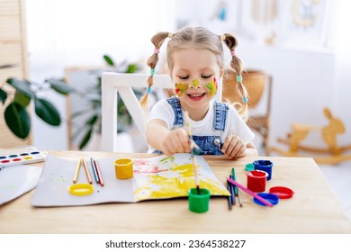 a little child girl paints a drawing in an album and smiles with a dirty face and hands, a happy child draws a picture at home at the table. - Powered by Shutterstock