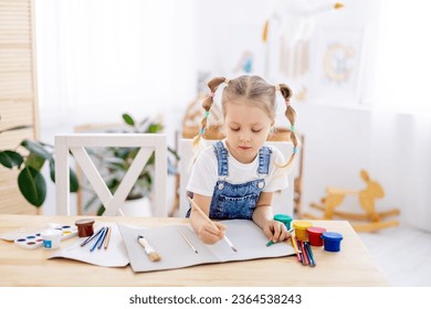 a little child girl paints a drawing with a brush in an album and smiles, a happy child draws a gouache picture at home at the table. - Powered by Shutterstock