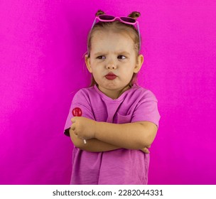 Little child girl pouted with a lollipop in her hands. Child girl cutely pouted her lips and folded her arms on her chest on a pink background. The concept of a funny little girl with a lollipop.