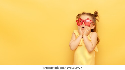 Little child girl in striped swimsuit and red funny summer sunglasses surprised expression looks at camera posing on yellow background, studio portrait.Advertising of children's products and sale