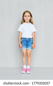 Little Child Girl In White T-shirt, Jeans Shorts And Pink Sneakers Standing Straight Near The Wall