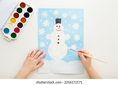 Little child hand holding paint brush   drawing snowman   snowflakes light blue paper white table background  Closeup  Point view shot  Toddler development 
