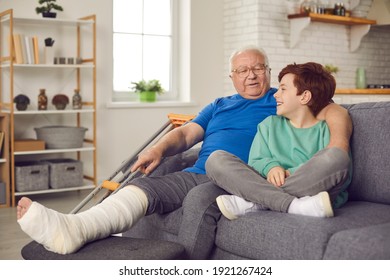 Little Child Helps, Supports And Takes Good Care Of His Grandpa With Injury. Happy Smiling Grandson And His Grandfather With Broken Leg In Cast Sitting On Sofa, Hugging And Talking