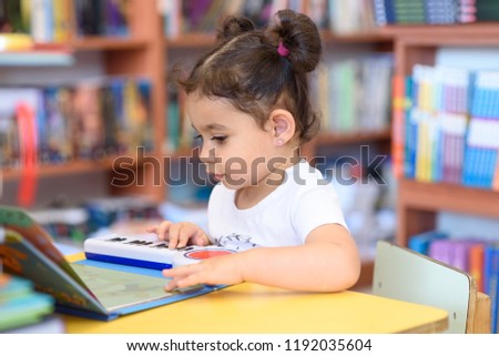 Little Child Indoors In Front Of Books. Cute Young Toddler Sitting On A Chair Near Table and play with toy piano. Kid in a bookstore, surrounded by colorful books. Happy multirace girl play the piano.