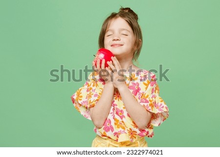 Little child kid girl 6-7 years old wear casual clothes have fun hold in hand red apple close eyes isolated on plain pastel green background studio portrait. Mother's Day love family lifestyle concept