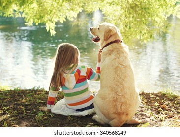 Little Child With Labrador Retriever Dog Sitting In Sunny Summer Park Near Water