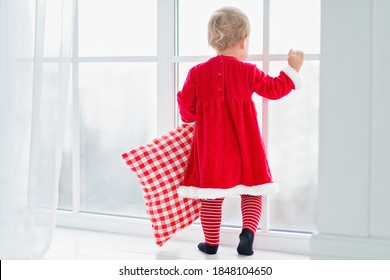 Little Child At Large Window, Wearing A Santa Style Dress And Hat Waiting For Christmas, Santa Claus. Holiday Concept,