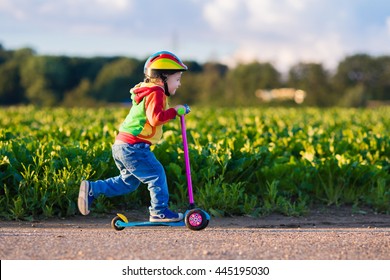 Little child learning to ride a scooter in a city park on sunny summer day. Cute preschooler boy in safety helmet riding a roller. Kids play outdoors. Active leisure and outdoor sport for children.