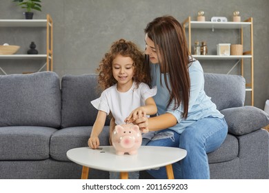 Little Child Learning The Value Of Money. Happy Young Mother And Cute Daughter Who Plan On Saving Up Some Money Sitting On Sofa At Home And Putting Coins In Piggy Bank. Concept Of Family Finance